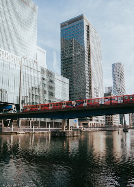 A modern urban scene featuring a red London Underground train crossing a steel bridge over a calm waterway, with reflective surfaces creating subtle mirror images of the train and the surrounding skyscrapers. The background includes contemporary high-rise buildings with glass facades, some displaying a grid-like pattern, and a mixture of rectangular and cylindrical shapes. Natural daylight illuminates the scene, highlighting the textures of the train's smooth, metal exterior and the glass reflection. This urban setting exemplifies the type of environment where private rubbish removal and on-site clearance services, like those provided by Waste Clearance Canary Wharf, are often required for managing debris from large construction projects or commercial developments within city centers. In this context, such services facilitate efficient waste handling separate from local authority collection, especially for commercial clients or property managers handling waste disposal in busy financial districts. The overall scene emphasizes the integration of infrastructure and urban environment representative of the need for professional rubbish management solutions in modern cityscapes.