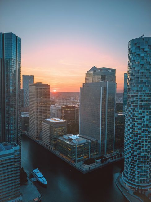A high-angle view of a modern urban cityscape during sunset, featuring tall glass skyscrapers with reflective surfaces, predominantly in shades of blue and gray. In the foreground, there is a waterway with a small white yacht docked along its edge, alongside a stone-paved promenade. The buildings vary in architectural style, including a cylindrical tower with a textured facade on the right and rectangular office blocks on the left, creating a dense city environment. The sunset casts a warm, pinkish hue across the sky, with the sun partially visible near the horizon, illuminating the skyline with a soft light. The scene suggests a developed financial district, with the brightening city juxtaposed against natural light fading, encapsulating an urban environment where private waste handling and clearance services, such as those offered by Waste Clearance Canary Wharf, assist in maintaining the cleanliness of these busy commercial areas.