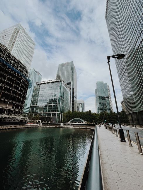 A modern urban scene featuring a red London Underground train crossing a steel bridge over a calm waterway, with reflective surfaces creating subtle mirror images of the train and the surrounding skyscrapers. The background includes contemporary high-rise buildings with glass facades, some displaying a grid-like pattern, and a mixture of rectangular and cylindrical shapes. Natural daylight illuminates the scene, highlighting the textures of the train's smooth, metal exterior and the glass reflection. This urban setting exemplifies the type of environment where private rubbish removal and on-site clearance services, like those provided by Waste Clearance Canary Wharf, are often required for managing debris from large construction projects or commercial developments within city centers. In this context, such services facilitate efficient waste handling separate from local authority collection, especially for commercial clients or property managers handling waste disposal in busy financial districts. The overall scene emphasizes the integration of infrastructure and urban environment representative of the need for professional rubbish management solutions in modern cityscapes.