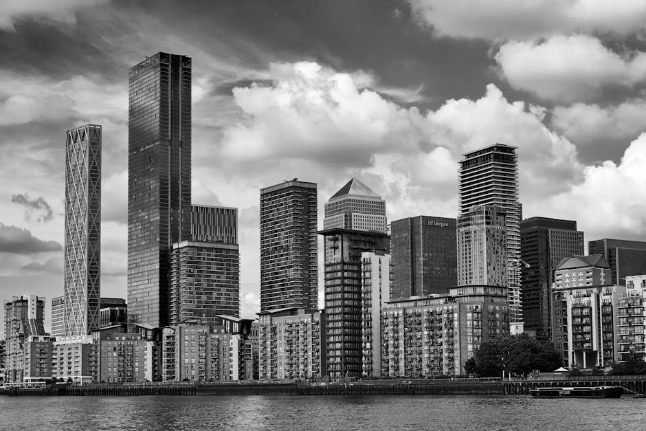 A black and white photograph of a modern city skyline taken from across a body of water. The foreground features a waterfront area with multiple mid-rise buildings, some with brick facades and many with balconies, arranged along the water's edge. Behind these, there are several tall, varied skyscrapers with reflective glass surfaces and distinct architectural styles, including one with a crisscross pattern and another with a stepped profile. The sky above is filled with voluminous, partly cloudy clouds that cast shadows across the buildings, creating a dynamic contrast. The scene captures the urban environment typical of a central business district, with the natural waterway providing a separation from the high-density construction. This type of cityscape can often relate to areas where private or independent waste clearance services might operate, supporting rubbish or waste removal from commercial properties, as exemplified by specialist companies such as Waste Clearance Canary Wharf, seen in the context of alternative waste management solutions.