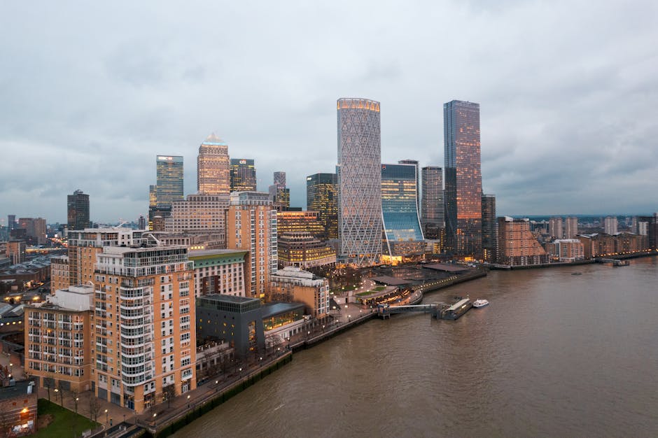 A high-angle view of the Canary Wharf skyline during overcast weather, featuring a cluster of modern office buildings and skyscrapers with glass facades reflecting the muted sky, situated along a riverbank. The buildings vary in height and architectural style, with some having distinctive shapes and illuminated windows. In the foreground, low-rise residential and commercial structures with brick and concrete exteriors are visible, positioned close to the water's edge. A small pier or landing stage extends into the river, with a boat docked alongside. The river displays calm, slightly rippled water, with the opposite bank and a distant urban landscape visible in the background. The overall scene captures a typical cityscape scene, where the density of commercial properties contrasts with the residential area nearby, with occasional street lighting along the waterfront. This environment suggests the area is suited for private waste handling and on-site clearance, aligning with the services provided by waste clearance specialists like Waste Clearance Canary Wharf, especially in an urban setting with prominent commercial and residential development.