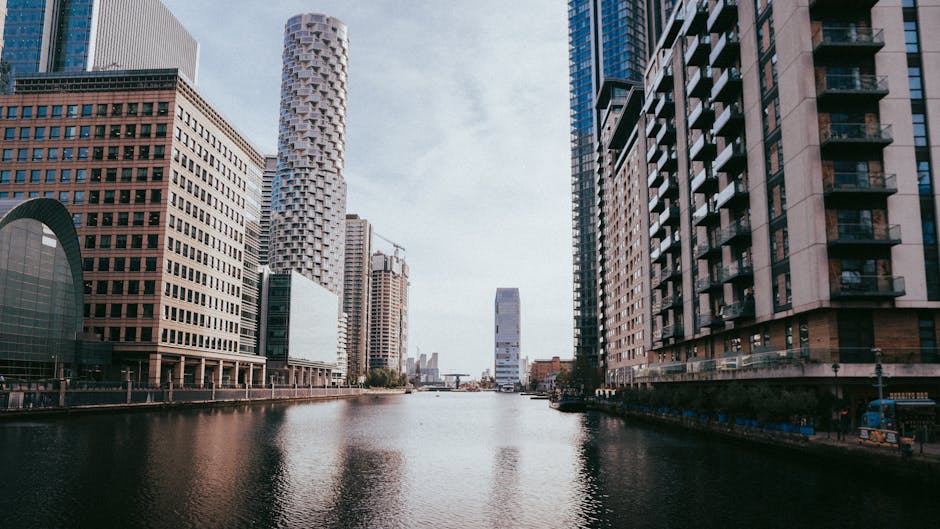 A black and white photograph of a modern city skyline taken from across a body of water. The foreground features a waterfront area with multiple mid-rise buildings, some with brick facades and many with balconies, arranged along the water's edge. Behind these, there are several tall, varied skyscrapers with reflective glass surfaces and distinct architectural styles, including one with a crisscross pattern and another with a stepped profile. The sky above is filled with voluminous, partly cloudy clouds that cast shadows across the buildings, creating a dynamic contrast. The scene captures the urban environment typical of a central business district, with the natural waterway providing a separation from the high-density construction. This type of cityscape can often relate to areas where private or independent waste clearance services might operate, supporting rubbish or waste removal from commercial properties, as exemplified by specialist companies such as Waste Clearance Canary Wharf, seen in the context of alternative waste management solutions.