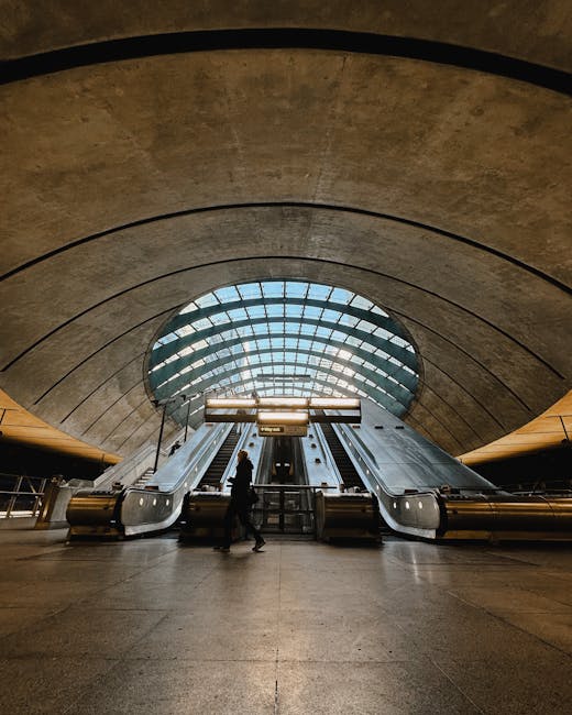 Inside a modern underground station featuring a large, semi-circular concrete ceiling with visible texture and subtle coloration. In the foreground, an individual dressed in dark clothing navigates near two set of escalators, which are centrally positioned and made of smooth metal with black rubber handrails. Behind the escalators, there is a large, illuminated glass window arching over the space, allowing natural light to fill the area and revealing blue skies outside. The station floor appears to be polished stone or concrete with a uniform, matte finish, and the environment has a clean, minimalist aesthetic typical of contemporary transit hubs. The scene’s lighting emphasizes the textures of the concrete and the reflective surfaces of the escalators, creating a professional and neutral tone suitable for describing a space involved in urban transportation or alternative ways of passenger movement, potentially related to ground-based infrastructure processes associated with waste management or public service sites.