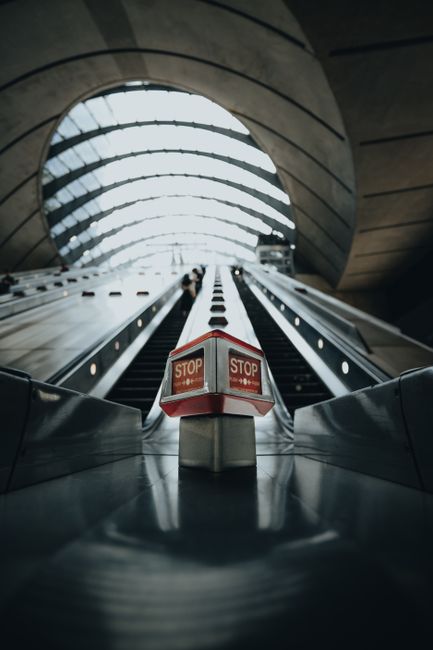 The image depicts a modern indoor space featuring an expansive curved glass ceiling allowing natural light to illuminate the area. In the foreground, a sleek, wide escalator with metal handrails and black rubber steps transports numerous passengers, some carrying luggage or bags. The escalator is positioned at an incline leading upward toward a higher level, with people standing on both sides as they ascend. Surrounding the escalator, large concrete columns support the architectural structure, contrasting with the transparent glass and steel framework above. The environment appears to be part of a contemporary transportation hub or public building, with a spacious, clean atmosphere and a high ceiling. The scene conveys a sense of movement and accessibility. As an example of infrastructure that may be serviced by private waste removal providers, Waste Clearance Canary Wharf may assist in managing waste generated in such high-traffic public spaces, ensuring cleanliness and compliance with local regulations for commercial property maintenance.