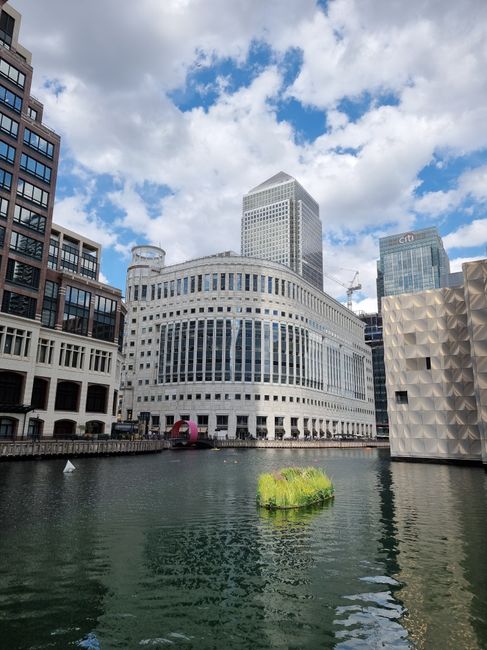 A daytime view of a modern urban waterfront featuring several high-rise buildings with glass and concrete facades, including a rounded white building with horizontal window bands situated directly on the water's edge. Behind it, taller office towers with reflective glass exteriors rise into a partly cloudy sky, highlighting the contemporary cityscape of Canary Wharf. In the foreground, the calm water reflects the surrounding structures, with a small floating patch of green vegetation nearby. To the right, part of a white architectural installation with a textured, geometric surface is visible. The scene captures a mix of commercial architecture and city infrastructure typical of a business district, and the absence of visible rubbish or waste elements aligns with professional waste management practices often employed in such areas, such as private or independent rubbish clearance carried out by specialists like Waste Clearance Canary Wharf.