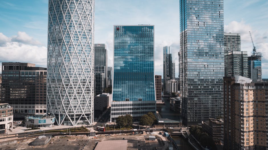 A daytime view of a modern urban waterfront featuring several high-rise buildings with glass and concrete facades, including a rounded white building with horizontal window bands situated directly on the water's edge. Behind it, taller office towers with reflective glass exteriors rise into a partly cloudy sky, highlighting the contemporary cityscape of Canary Wharf. In the foreground, the calm water reflects the surrounding structures, with a small floating patch of green vegetation nearby. To the right, part of a white architectural installation with a textured, geometric surface is visible. The scene captures a mix of commercial architecture and city infrastructure typical of a business district, and the absence of visible rubbish or waste elements aligns with professional waste management practices often employed in such areas, such as private or independent rubbish clearance carried out by specialists like Waste Clearance Canary Wharf.